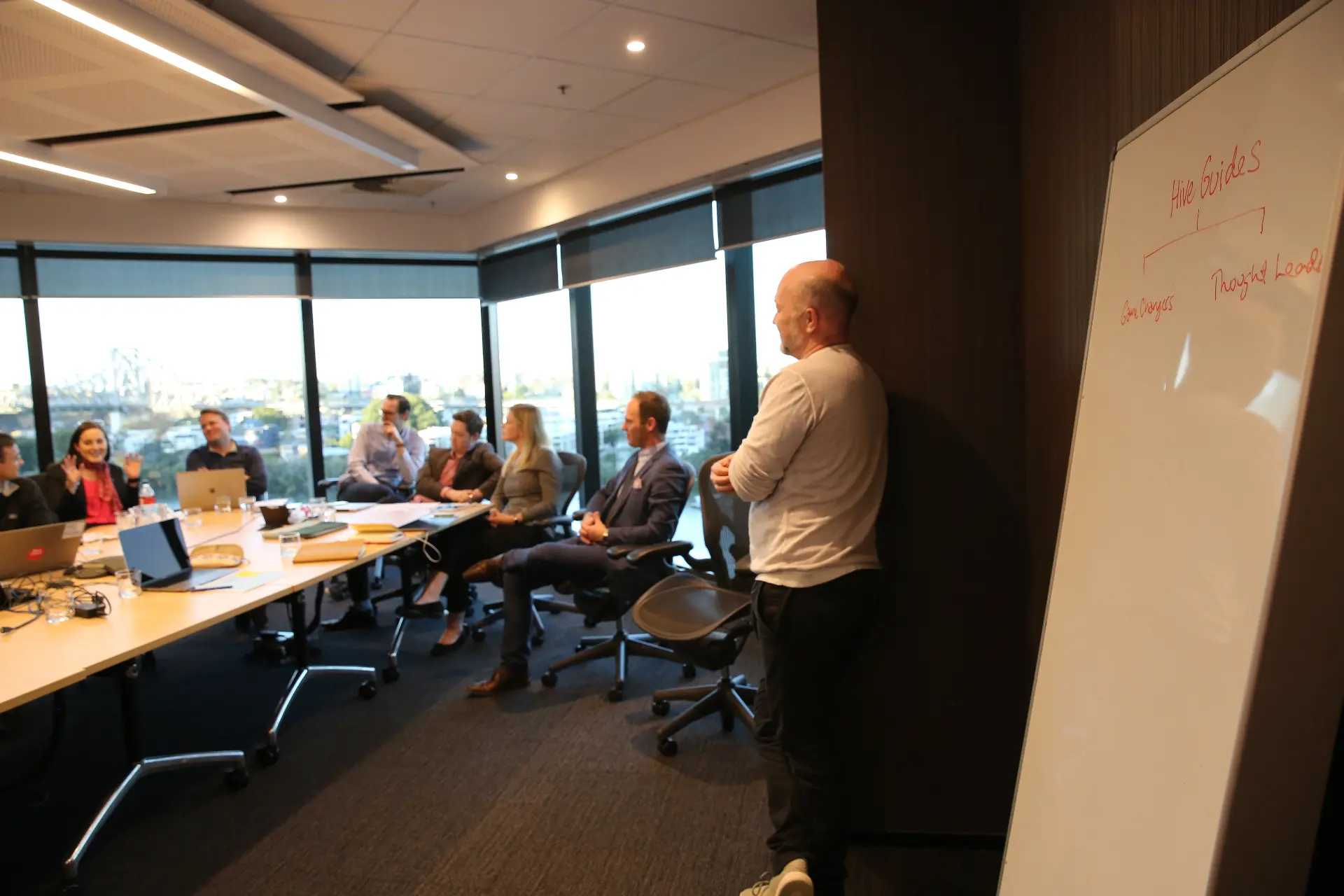 A facilitator standing at a whiteboard headed Hive Guides, leading a boardroom of eight people seated along a window with the Story Bridge behind them.
