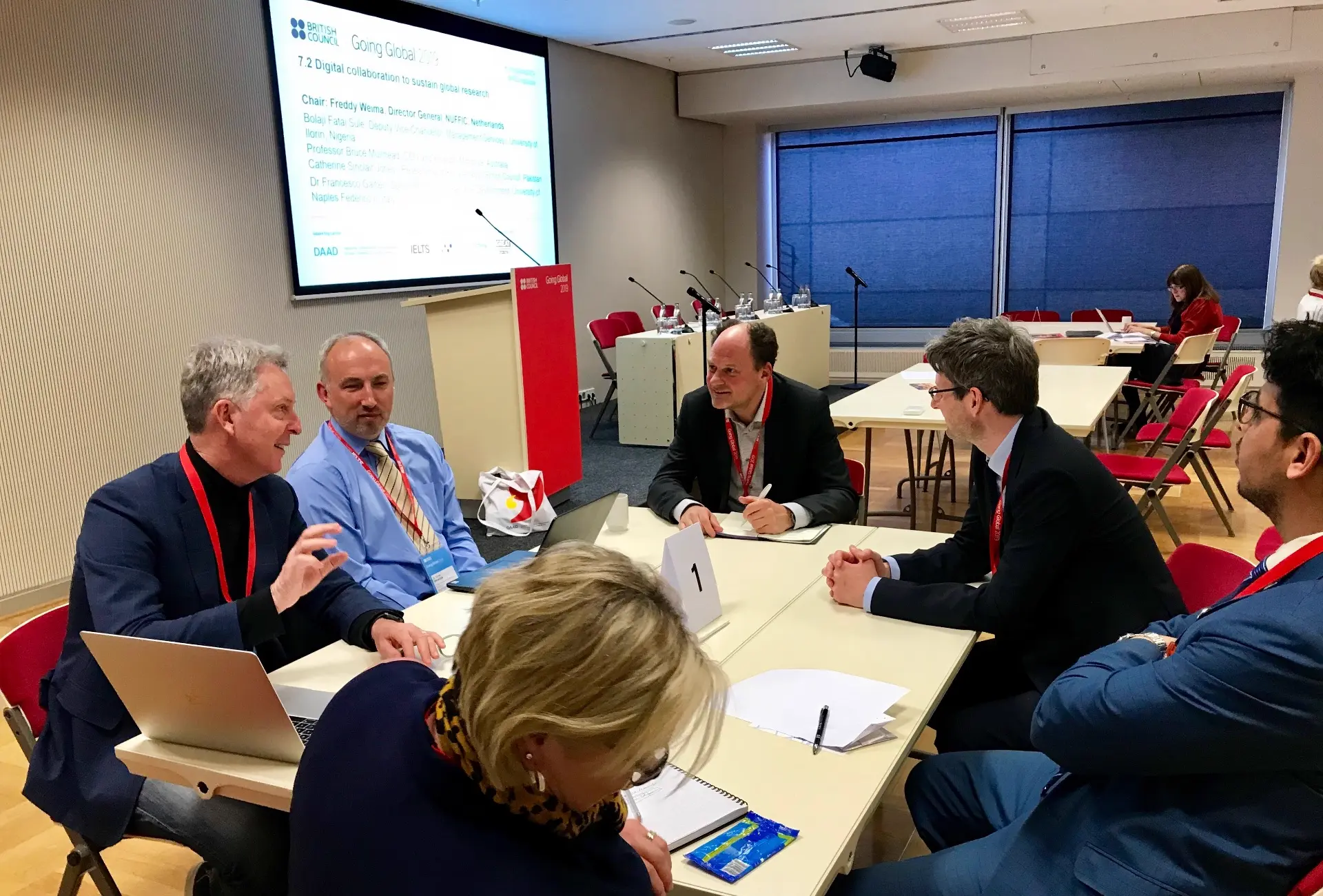 Six delegates around a small conference table in a facilitated working session, one of them talking while the others take notes.