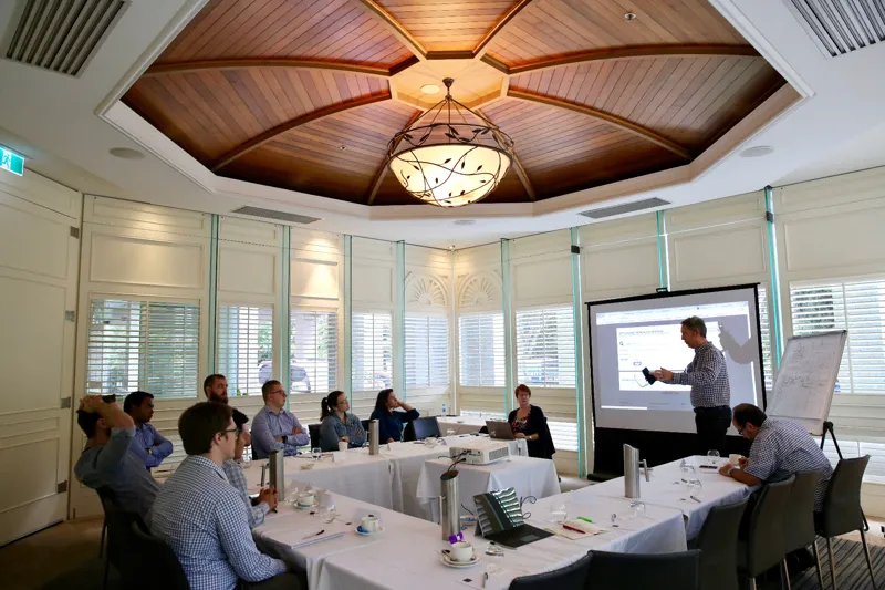 A facilitator presenting to a dozen seated participants at a workshop, working from a projected screen and a flip chart.